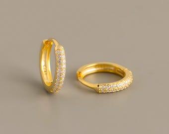 May include: A pair of gold hoop earrings featuring a hinged closure. Each hoop is adorned with a single row of small, clear, round-cut stones embedded along the outer surface, creating a sparkling effect against the polished gold metal finish.
