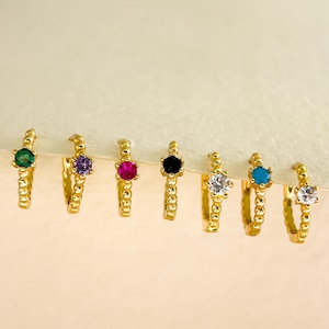 May include: A set of eight gold hoop earrings with small colourful gemstones. The gemstones are green, purple, pink, black, white, turquoise, and two white gemstones.