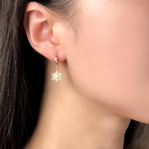 May include: A gold hoop earring featuring a dangling starburst charm. The starburst is crafted from gold metal and is encrusted with small, clear, sparkling stones arranged in a radiating pattern that extends from a central circular point.