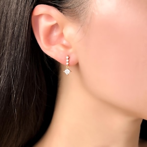 May include: A gold drop earring featuring a vertical row of three small round clear stones leading to a single square-cut clear stone at the base. The earring is suspended from a gold post that secures directly into the earlobe, creating a delicate and refined appearance with a polished metallic finish.