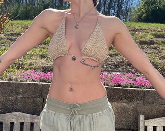 May include: A woman wearing a beige crocheted bikini top with a silver chain and a small pendant. She has a tattoo on her left side that says "Forever in my heart".