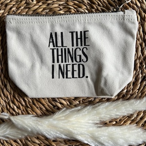 Könnte beinhalten: Eine weiße Canvas-Kosmetiktasche mit schwarzem Text, der "All the things I need." lautet.