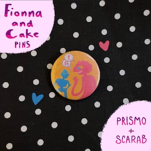 Może przedstawiać: Okrągła przypinka z kreskówkową ilustracją dwóch postaci w kolorze niebieskim i różowym na pomarańczowym tle. Przypinka jest oznaczona "Fionna and Cake PINS" i "Prismo + Scarab" różowym tekstem. Tło jest czarne z białymi kropkami.