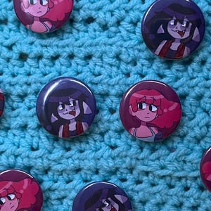 May include: Six colourful round buttons featuring cartoon characters. The buttons depict two different characters, one with pink hair and one with purple hair. The characters are wearing different outfits and have different expressions. The buttons are displayed on a blue crocheted surface.