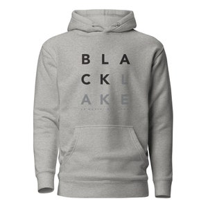 Könnte beinhalten: Ein hellgrauer Kapuzenpullover mit einer Vordertasche und einer Kapuze. Das Wort "BLACK LAKE" ist vertikal in schwarzen und grauen Buchstaben aufgedruckt. Darunter steht der Satz "UP NORTH, USA".