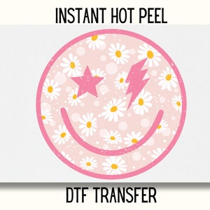 May include: Pink smiley face with a white daisy pattern and a pink lightning bolt and star. The text "INSTANT HOT PEEL" and "DTF TRANSFER" is printed below the image.