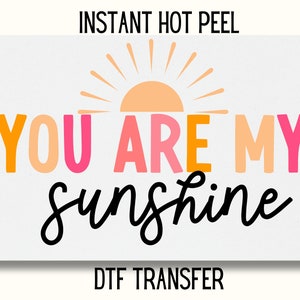 May include: A white background with a black and white graphic that says "You are my sunshine" with a stylized orange sun above the text. The text is in a rainbow of colors. The words "Instant Hot Peel DTF Transfer" are at the top and bottom of the image.