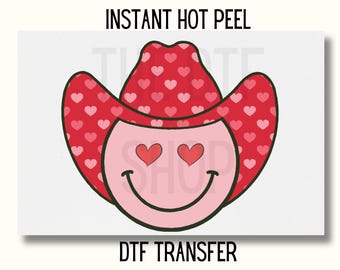 Puede incluir: Una cara sonriente rosa con un sombrero de vaquero rojo con un patrón de corazón. La cara sonriente tiene corazones por ojos y una gran sonrisa. El texto "INSTANT HOT PEEL" y "DTF TRANSFER" están en la parte superior e inferior de la imagen.