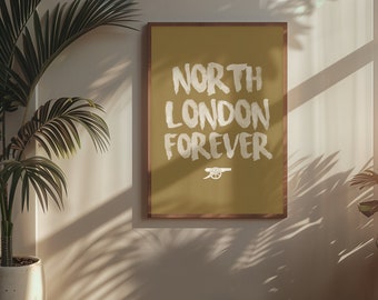 May include: A framed poster with the words "NORTH LONDON FOREVER" in white brush lettering. The poster has a gold background and a brown wooden frame. A small cannon logo is at the bottom. The poster is hanging on a white wall.