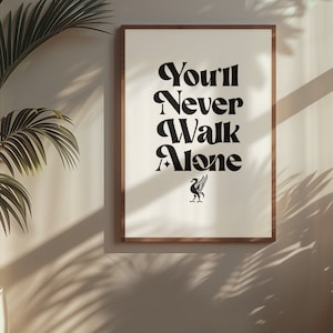 Op de afbeelding: Een ingelijst print met de tekst "You'll Never Walk Alone" in een retro lettertype. Onder de tekst staat een zwart en rood vogellogo.