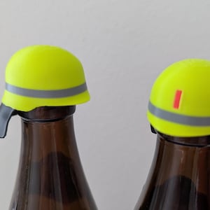 K&ouml;nnte beinhalten: Zwei gelbe Flaschenverschl&uuml;sse in Form von Bauhelmen mit einem grauen Streifen und einem roten Streifen. Die Verschl&uuml;sse befinden sich auf braunen Glasflaschen.