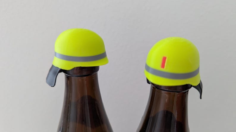 K&ouml;nnte beinhalten: Zwei gelbe Flaschenverschl&uuml;sse in Form von Bauhelmen mit einem grauen Streifen und einem roten Streifen. Die Verschl&uuml;sse befinden sich auf braunen Glasflaschen.