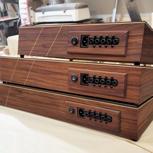 May include: Three stacked, dark wood audio equipment units with gold trim. Each unit has a power inlet, power switch, and multiple audio jacks. The units are arranged in a pyramid shape, with the largest at the bottom and the smallest on top.
