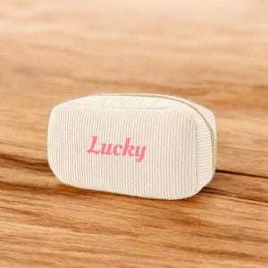 May include: A cream-colored corduroy pencil case with the word "Lucky" in pink script. The rectangular case has a rounded top and a gold zipper. The case is sitting on a wooden surface.