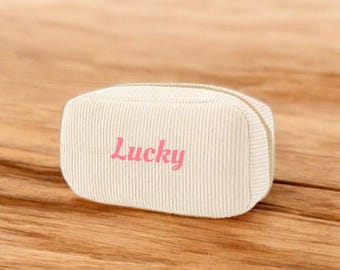 May include: A cream-colored corduroy pencil case with the word "Lucky" in pink script. The rectangular case has a rounded top and a gold zipper. The case is sitting on a wooden surface.