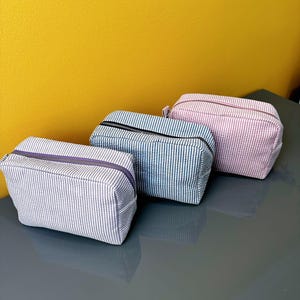 May include: Three rectangular cosmetic bags with vertical striped patterns. The bags are in shades of blue, pink, and gray, each with a contrasting zipper. The bags are arranged on a gray surface against a yellow background.