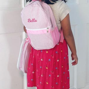 May include: A pink and white striped backpack with a pink zipper and the name "Bella" embroidered on the front. The backpack is being worn by a child wearing a red floral skirt.
