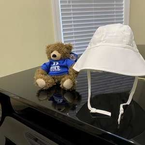 May include: A white sun hat with a wide brim and chin ties sits next to a brown teddy bear wearing a blue shirt with "DUKE" printed on it. The items are on a glossy black surface.