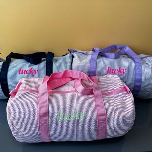 May include: Three seersucker duffel bags in blue, pink, and lavender, each with a matching handle and the word "lucky" embroidered on the side. The bags are on a dark gray surface.