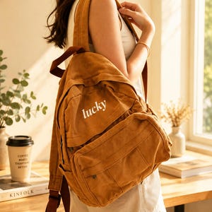 May include: A brown canvas backpack with the word "lucky" in white lettering. The backpack has multiple pockets and adjustable straps. The bag is worn over the shoulder. The overall style is casual and practical.
