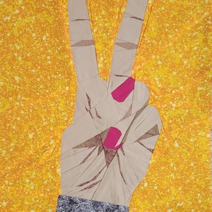 May include: A fabric art piece depicting a hand making the peace sign. The hand is beige with pink fingernails and a gray and black wristband. The background is yellow with white speckles.