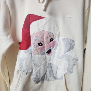 May include: A white hoodie with a patchwork Santa Claus design on the front. The Santa is made of different colored fabrics and has a red hat.