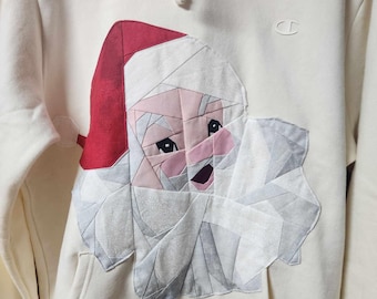 May include: A white hoodie with a patchwork Santa Claus design on the front. The Santa is made of different colored fabrics and has a red hat.