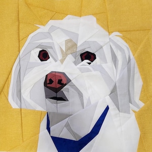Op de afbeelding: Een geometrisch stoffen kunstwerk met een witte hond met rode ogen en een roze neus. De hond draagt een blauwe kraag en staat tegen een gele achtergrond. Het kunstwerk is gemaakt van verschillende stoffen vormen.