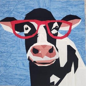 May include: A fabric quilt block featuring a cow's face with black and white patches, wearing red-rimmed glasses. The cow's nose is pink, and the background is light blue with a subtle pattern. The quilt block is square.