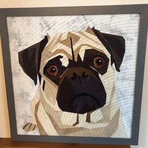 May include: A framed quilted artwork of a pug dog with brown eyes and a black nose. The dog is looking directly at the viewer. The background is a light gray with a subtle pattern.