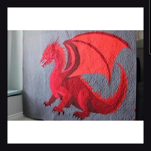 May include: A rectangular quilt featuring a large, vibrant red dragon with detailed wings and a long tail, centred against a textured grey background. The dragon is depicted in a side profile with sharp claws and a defined head, showcasing intricate stitching patterns throughout the fabric surface.
