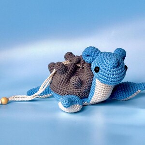 May include: A crocheted blue and gray turtle with a white belly and brown shell. The turtle has black eyes and a white string attached to its shell.