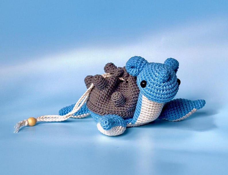 May include: A crocheted blue and gray turtle with a white belly and brown shell. The turtle has black eyes and a white string attached to its shell.