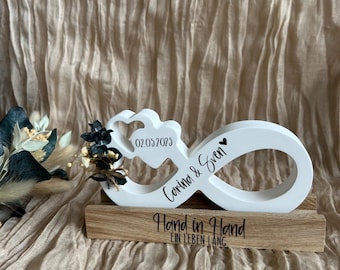 Puede incluir: Un símbolo de infinito de madera blanca con los nombres "Corina & Sven" y la fecha "02.05.2023". El símbolo de infinito está montado sobre una base de madera con las palabras "Hand in Hand" y "EIN LEBEN LANG" en escritura negra. Flores secas están unidas al lado izquierdo.