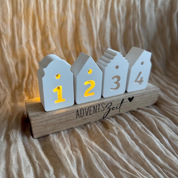 May include: A white wooden Advent calendar shaped like four small houses, each with a number from one to four. The numbers are illuminated with a warm yellow light. The base is a wooden plank with the text "ADVENTSZeit" and a heart.