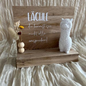 Könnte beinhalten: Ein Holzschild mit dem Text "Lächle du kannst sie nicht alle anspucken!" und einer weißen Keramik-Lama-Figur auf einem Regal.