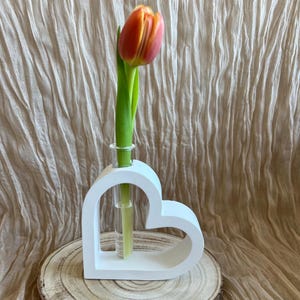 Könnte beinhalten: Weiße herzförmige Vase mit einer einzelnen orangefarbenen und gelben Tulpe in einem Glasröhrchen. Die Vase steht auf einer Holzoberfläche.
