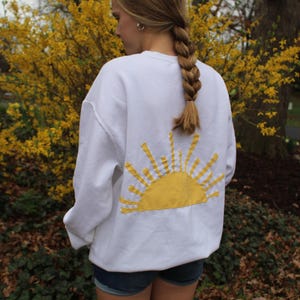 May include: White sweatshirt with a yellow embroidered sun design on the back. The sweatshirt is worn by a person with long brown hair.