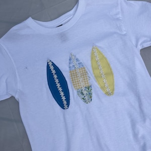 以下が含まれることがあります： 白いTシャツに、サーフボード型のアップリケが3つ付いています。アップリケは青、黄色、花柄とギンガムチェック柄です。各アップリケは白いデイジーチェーンで縁取られています。
