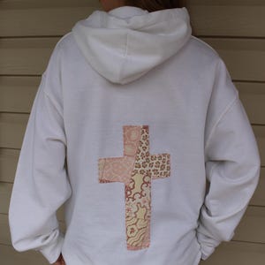 Può includere: Felpa bianca con un design a croce di patchwork sul retro. La croce è realizzata con diversi tessuti in tonalità di rosa, marrone e crema.