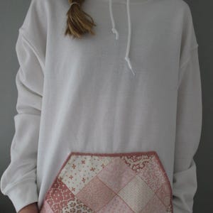 May include: White hoodie with a patchwork pocket. The pocket features a variety of pink and cream-coloured patterned fabrics, including floral and geometric designs. The hoodie has a drawstring hood and a kangaroo pocket.