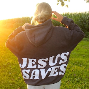 Può includere: Una felpa con cappuccio grigio scuro con la scritta "JESUS SAVES" a grandi lettere bianche sulla schiena. L'immagine è scattata all'aperto, con una persona che indossa la felpa, con un campo di erba verde e un tramonto luminoso sullo sfondo.