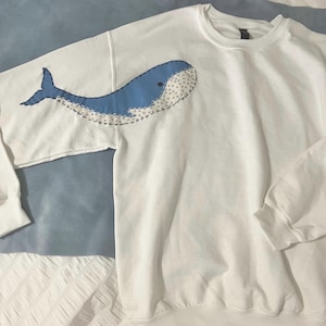 May include: White sweatshirt featuring a large blue whale applique on the left side. The whale design includes a light blue body, a dark blue tail, and a dotted pattern. The sweatshirt has long sleeves and a crew neck.