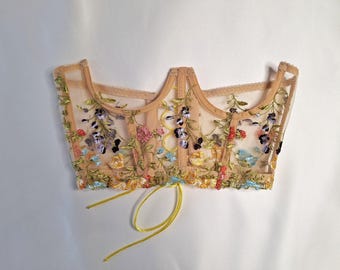 May include: A beige corset top with floral embroidery in various colors, including pink, blue, yellow, and purple. The corset has a sheer overlay and a yellow ribbon tie. The garment is designed with a sweetheart neckline and a structured bodice.