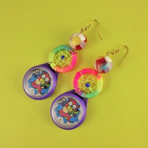 May include: A pair of colourful earrings featuring a rainbow dreamcatcher design and a purple maze-like charm with a cartoon character. The earrings are made with gold-coloured metal and have a beaded design.