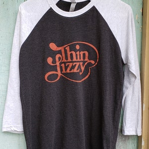 May include: A black and grey baseball tee shirt with a red and orange "Thin Lizzy" logo printed on the front.