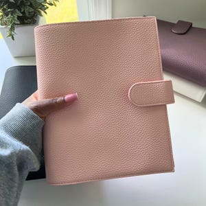 Op de afbeelding: Een lichtroze, getextureerde planner met een bijpassende sluiting. De planner wordt in een hand gehouden, met andere planners in zwart, wit en paars op de achtergrond. De planner heeft een afgeronde rug en een getextureerd oppervlak.
