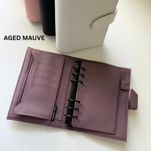 Op de afbeelding: Een open, verouderde mauvekleurige planner met een getextureerd oppervlak, zwarte ringbanden en meerdere binnenvakken. De planner is open, waardoor de binnenpagina's zichtbaar zijn. De tekst "AGED MAUVE" is zichtbaar.