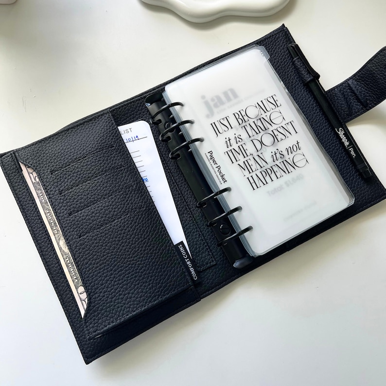 Op de afbeelding: Zwart leren planner met een zwarte pen en een doorzichtige papieren pocket. De papieren pocket heeft het woord "jan" en de tekst "JUST BECAUSE it is TAKING TIME, DOESN'T MEAN it's not HAPPENING". De planner is open en toont vakken en een lijst.