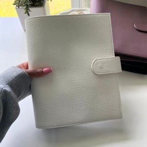 Op de afbeelding: Een witte, getextureerde leren planner met een gebogen klepsluiting. Op de achtergrond is een andere planner in mauve te zien. De planner wordt in een hand gehouden. De planner is neutraal van kleur en heeft een eenvoudig ontwerp.